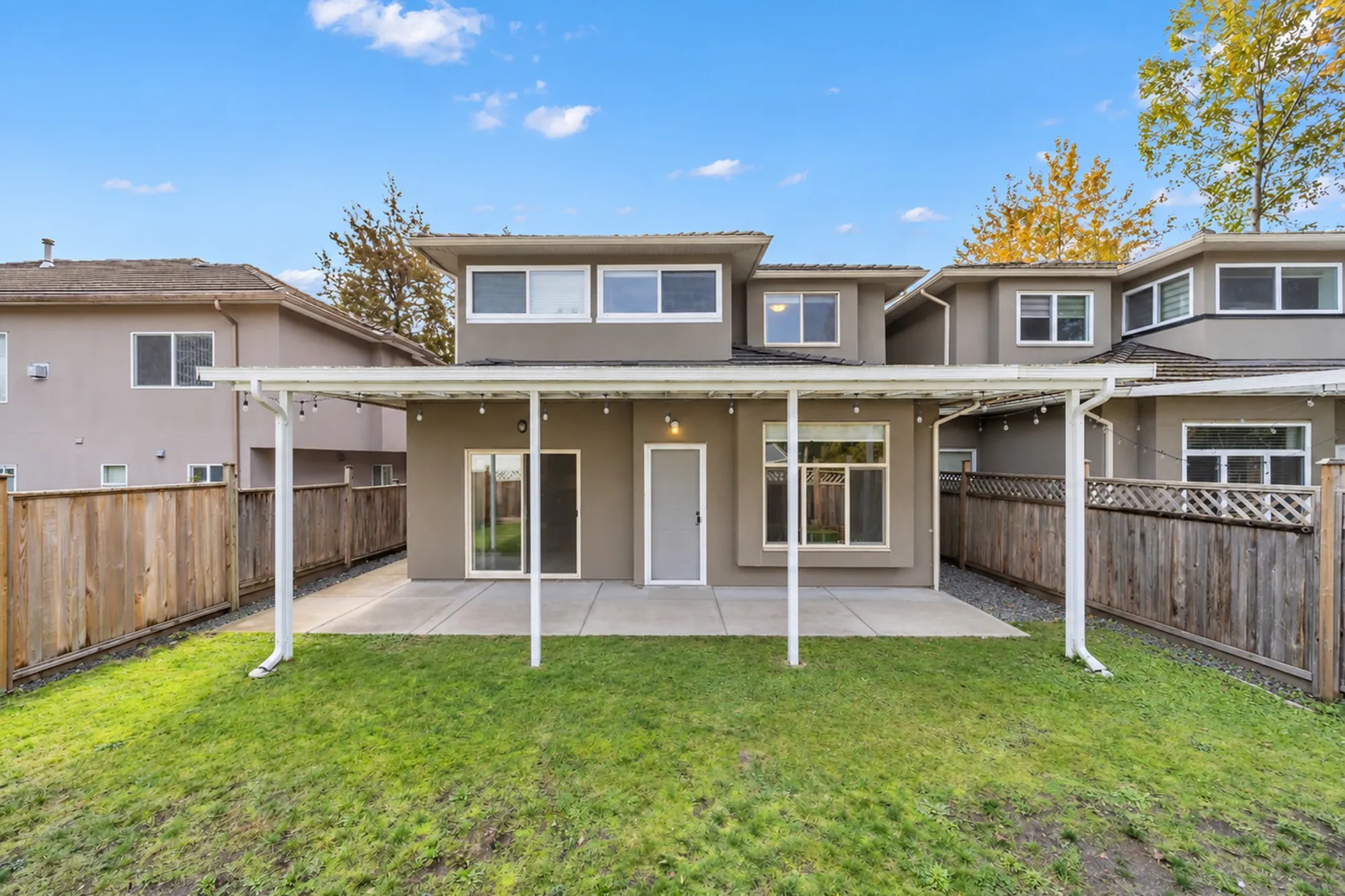 The back of the house: a covered patio on white posts across its width, a sliding door, a door and windows, wood fences each side, a lawn