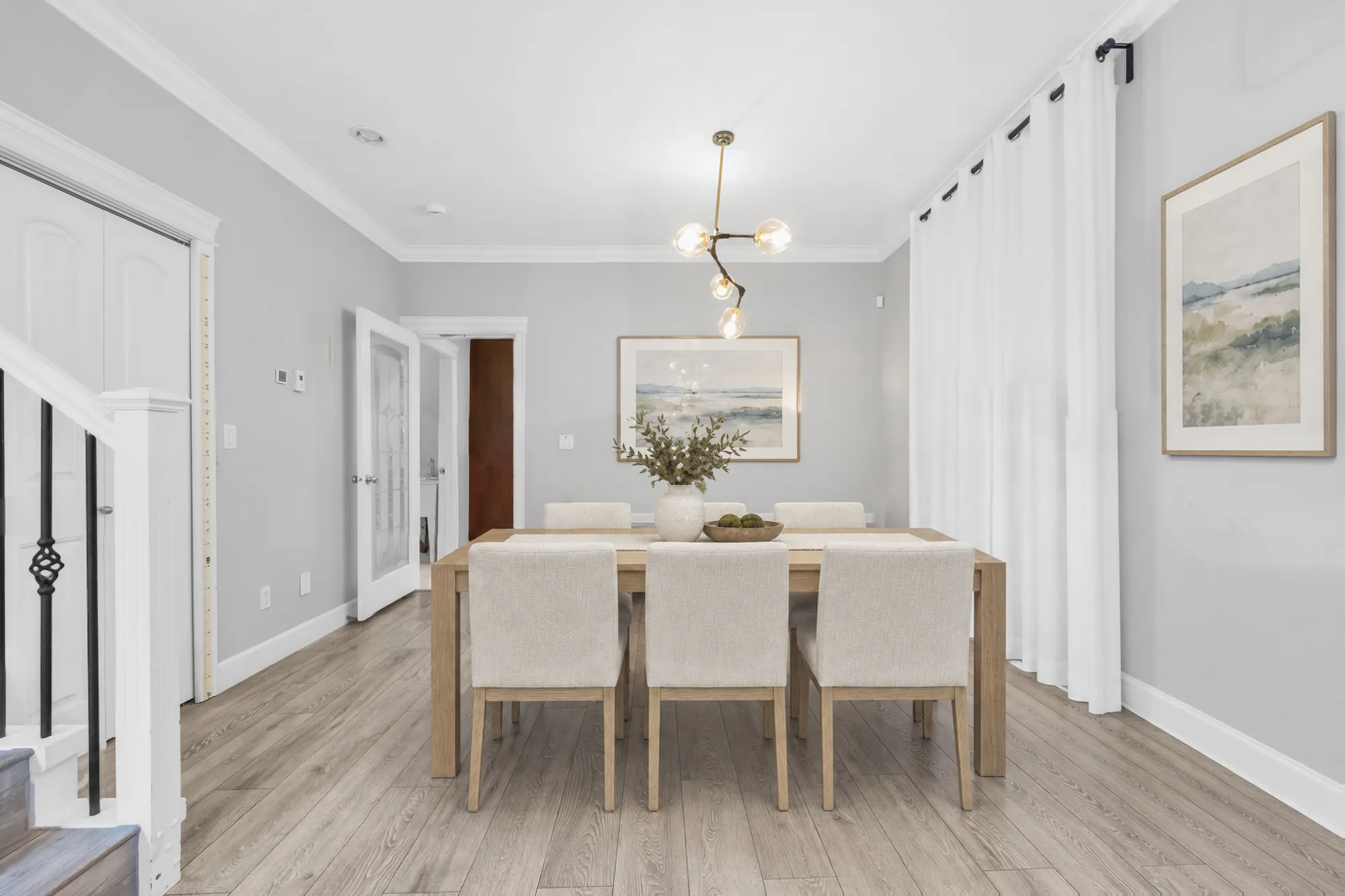 The dining room virtually staged: a pale wood table and upholstered chairs under a glass-globe pendant, sheer curtains, the stair at the left