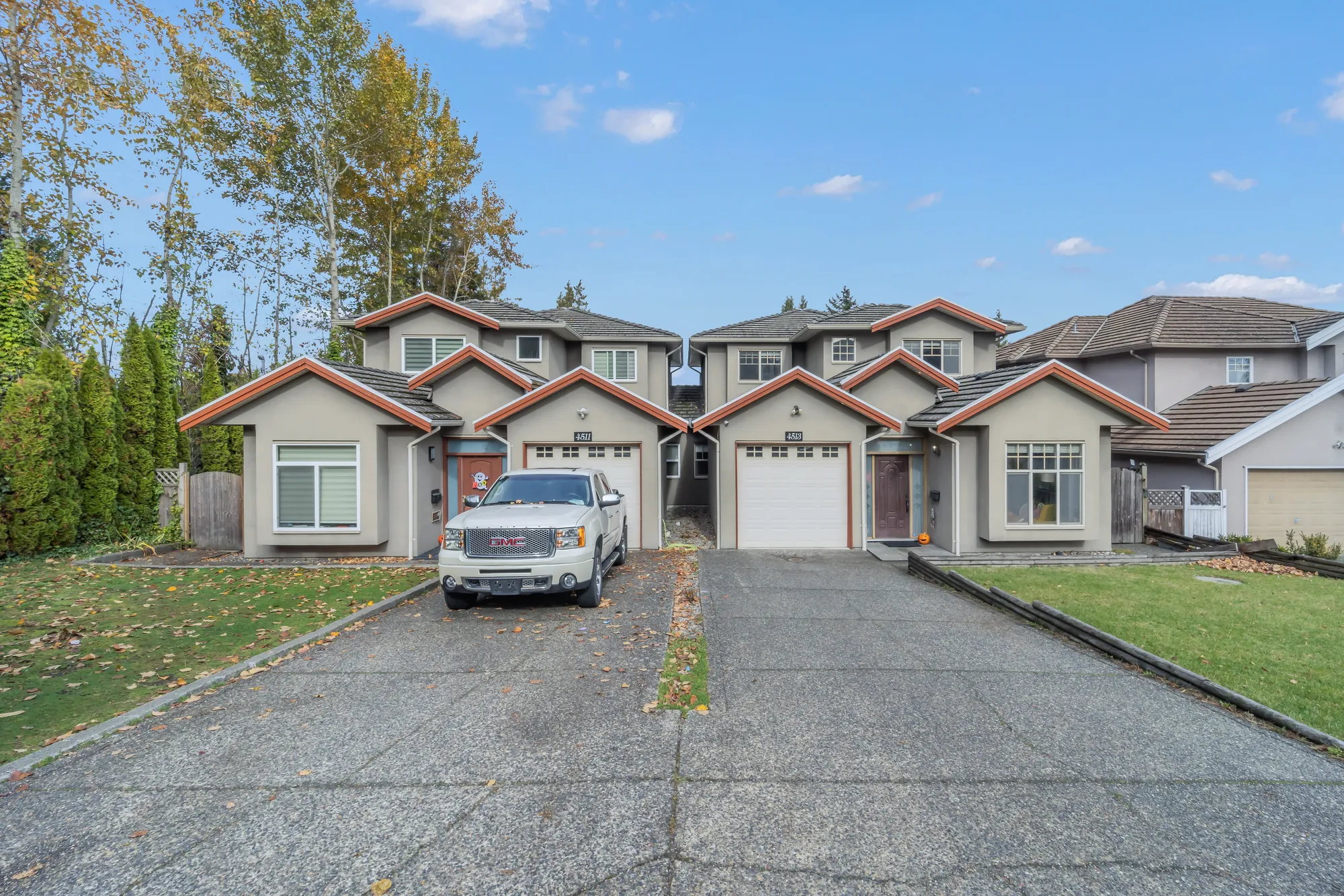 The duplex from the drive: two garage doors under gables trimmed in red-orange, a pickup on the drive, a lawn and a hedge of cedars