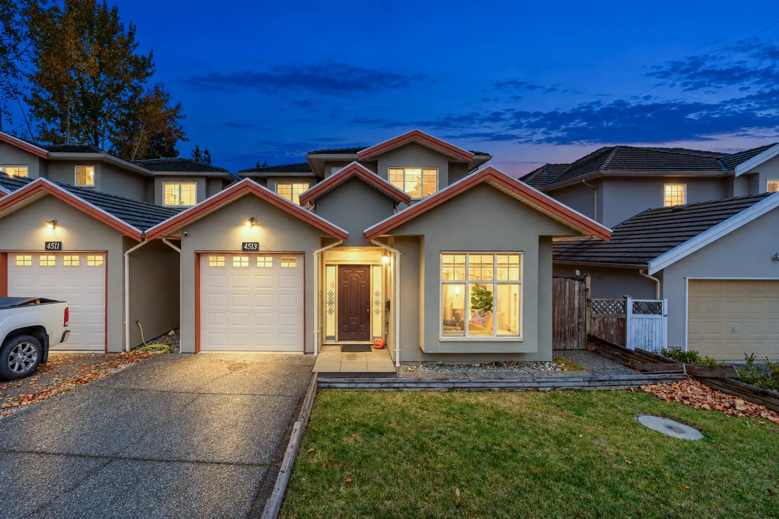 4513 Ellerton Court at dusk, an edited photograph: a grey stucco duplex with red-orange trim, the garage lights, the front door and the windows lit under a deep blue sky