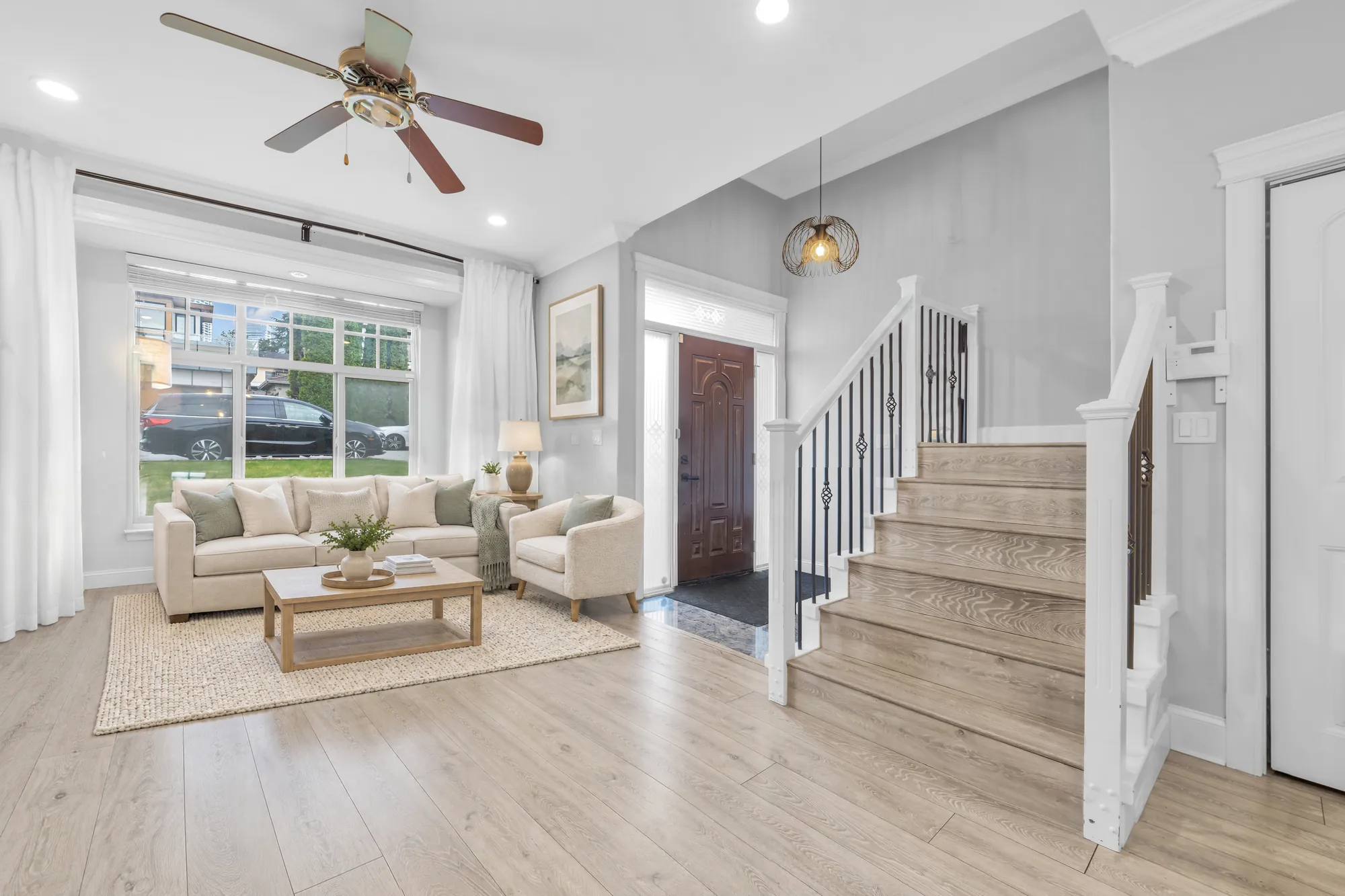 The living room virtually staged: a cream sofa and armchairs around a wood coffee table by the front window, a ceiling fan, the front door and the stair beside it