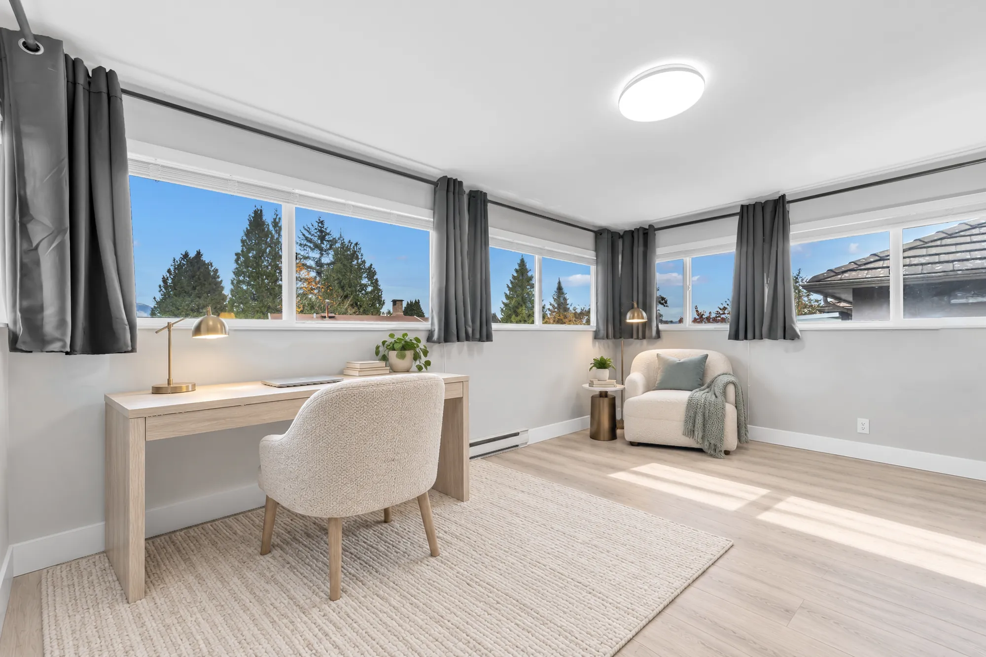 The office virtually staged: a pale wood desk and chair, an armchair, windows on two walls onto the treetops