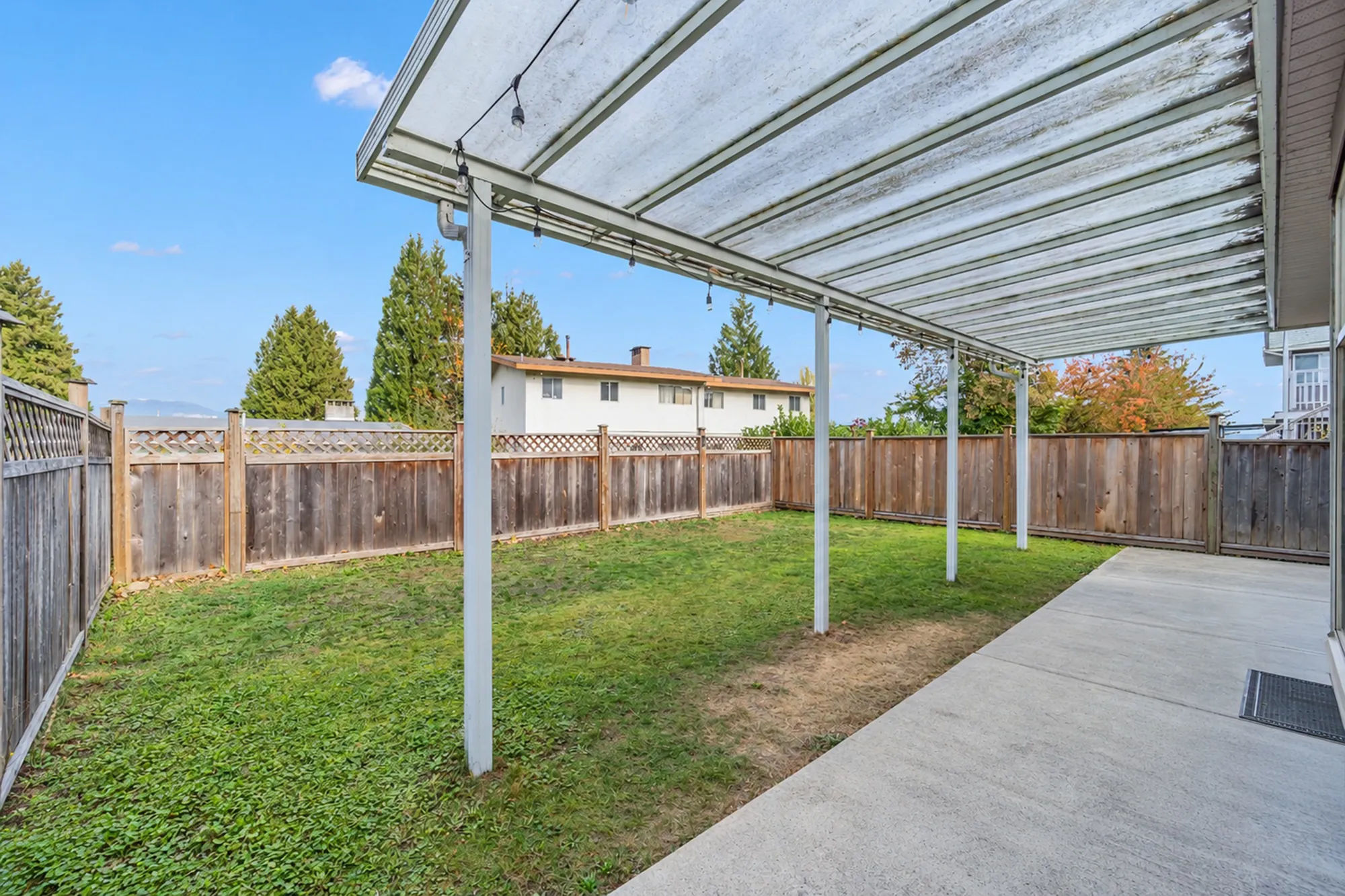 The covered patio: a translucent roof on white posts, string lights, a concrete slab, the fenced lawn beyond